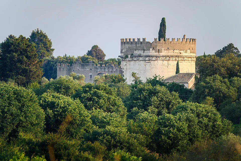 A pale round tower crowned with battlements rises above a dense mass of trees, with a tall cypress beside it, a lower crenellated building joined to it on the left and a tiled roof below it on the right.