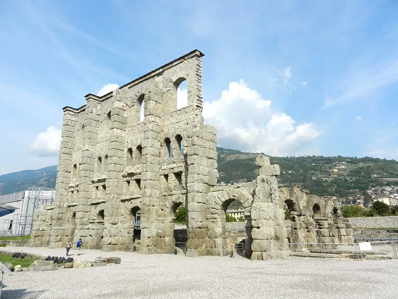 A tall, free-standing wall of grey stone blocks pierced by rows of arched and square openings, with broken piers and an arch beside it on a gravel area, and wooded mountainsides behind.