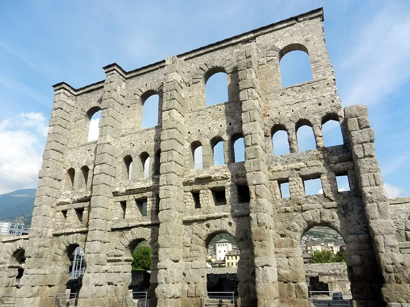 The face of the stone wall seen from close below, divided by projecting buttresses into bays, with arched entrances at the bottom and several rows of arched and square windows above.