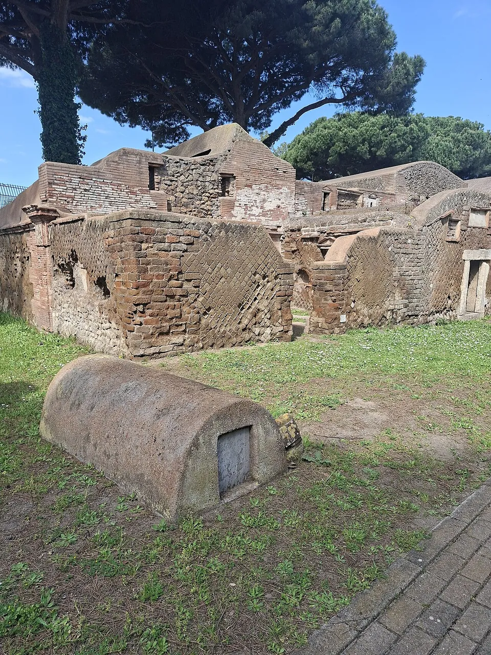 Brick tomb buildings with niches and broken vaults under umbrella pines, with a low half-cylinder tomb lying in the grass in front of them.