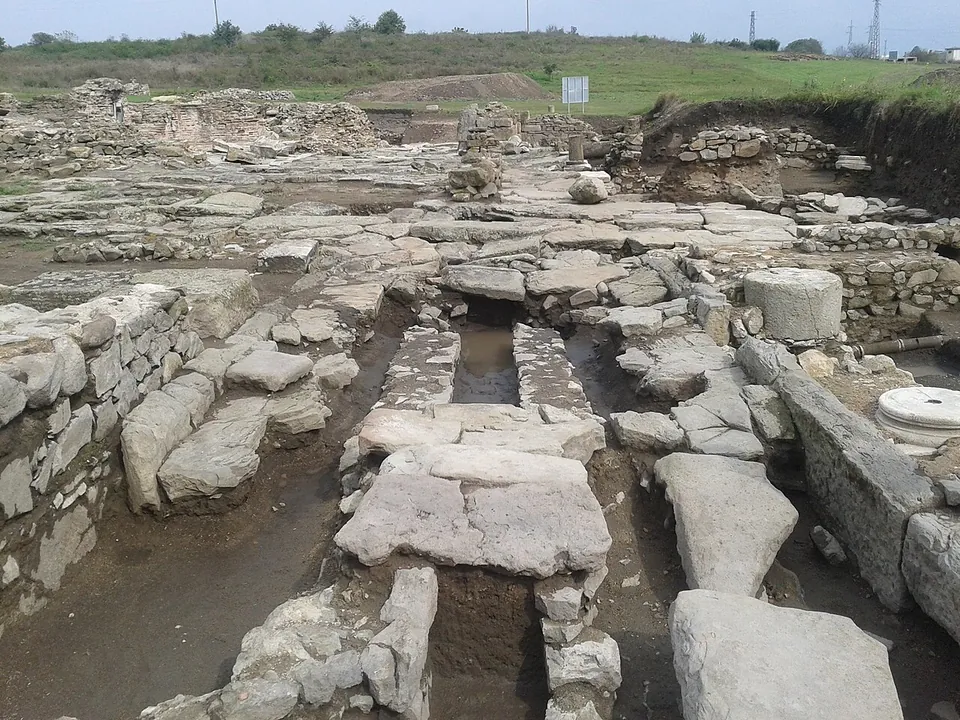 An excavation of stone foundations, paving slabs and stone-lined channels, with column bases and drums scattered among the walls and grassy ground behind.