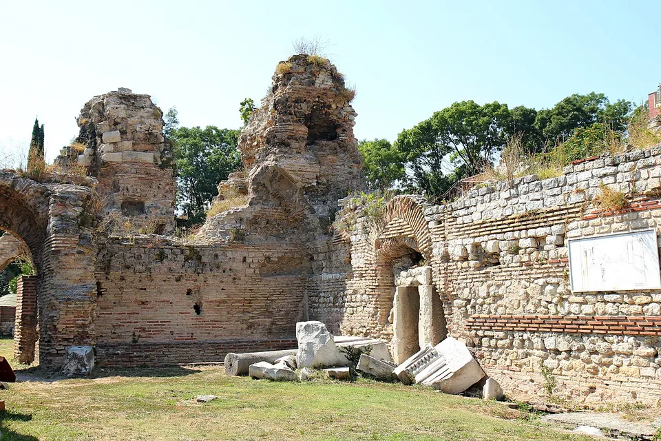 Ruined walls of brick and rough stone with several round-headed arches, broken high masonry above them, fallen pieces of white marble on the grass in front and an information board on the right.
