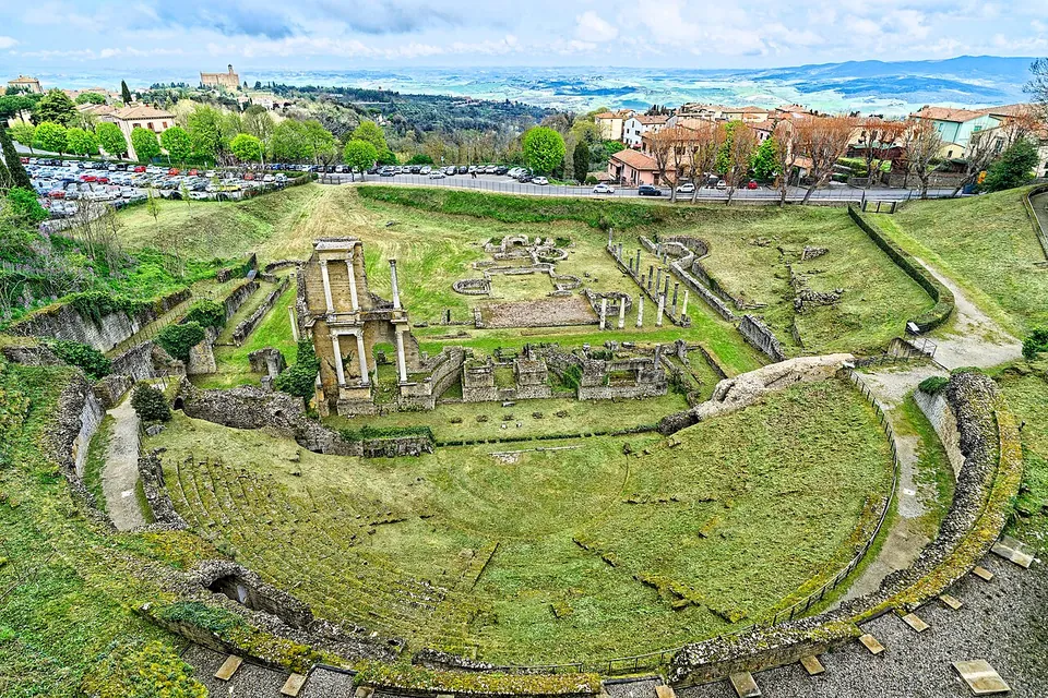 The Roman theatre from the slope above it, a grassy half-circle of seating, the standing fragment of the stage front and the excavated ground behind it, with a road, parked cars and houses at the edge of the town and hills on the horizon.