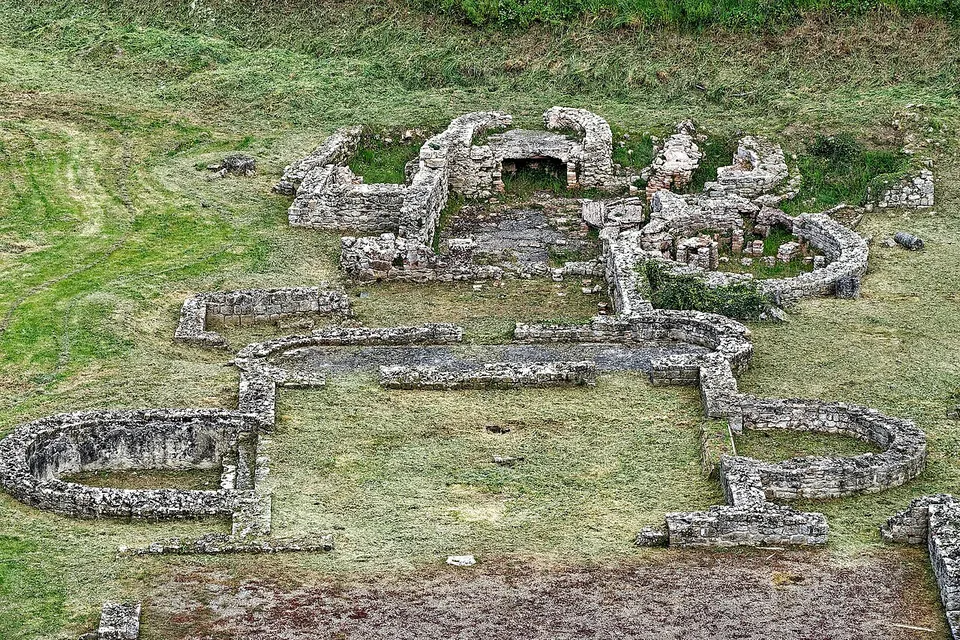 Low stone walls outlining a group of rooms on mown grass, several of them rounded at one end, seen from above.