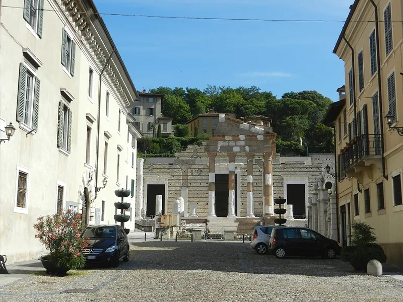 A cobbled square lined with pale four-storey houses and parked cars, closed at the far end by the columned front of the temple, with a wooded hill rising behind it.