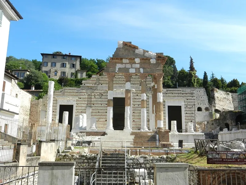 The front of a Roman temple on a high terrace, a porch of tall columns banded in pale stone and red brick under a broken triangular pediment, with a pale stone wall and three dark doorways behind it, a wide flight of steps in front and railings around excavated ground below.