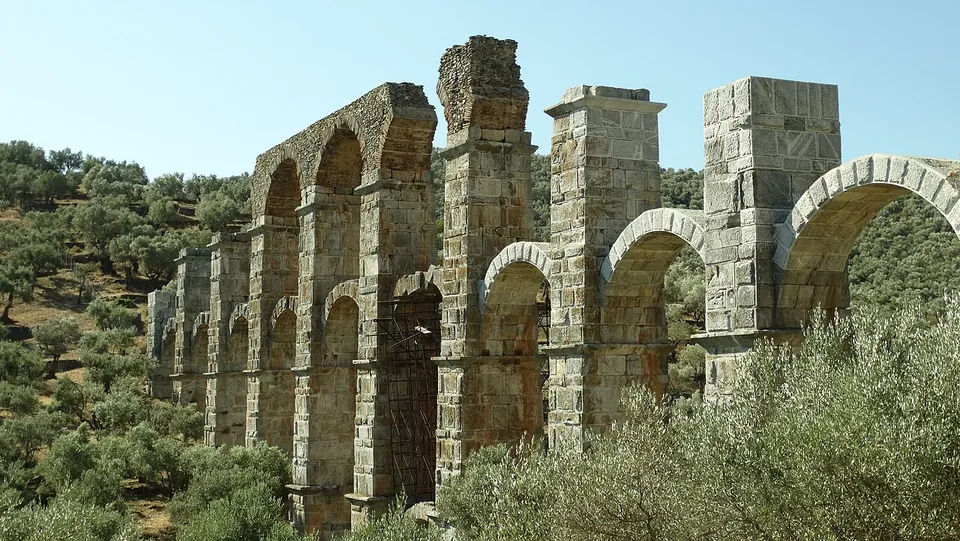 A tall arcade of grey stone piers carrying round arches in two tiers, partly broken off at the top, rising from olive groves on a hillside under a clear sky.