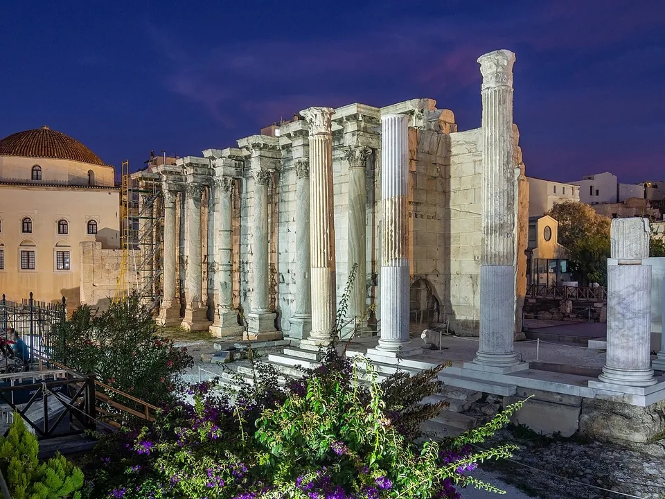 A tall wall of grey marble blocks lined with a row of Corinthian columns standing close against it, lit at dusk, with flowering bushes in front and a domed building to the left.