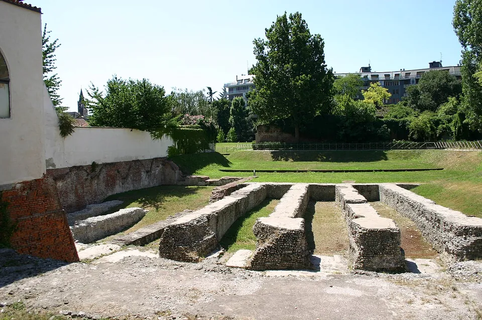 Low parallel walls of rough concrete and stone running out into a lawn from the white and brick wall of a building, with trees and blocks of flats beyond.