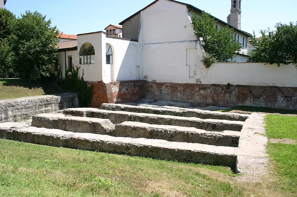 Stepped rows of low foundation walls in grass in front of a white building with an arched window and a brick base, a bell tower rising behind.