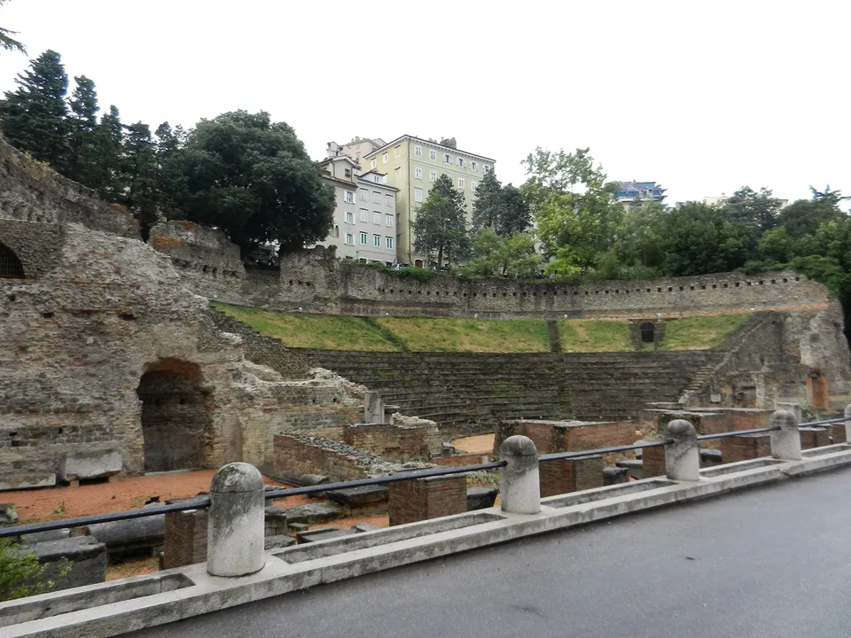 A pavement with a low metal railing and stone bollards, and beyond it, below street level, the ruins of the stage area with the curved seating and its high wall behind, under trees and houses.