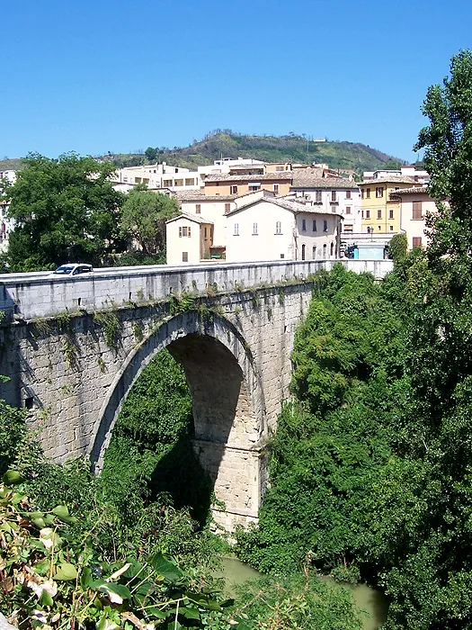 A single high round arch of pale squared stone blocks rises out of a deep, tree-filled gorge, carrying a road with a car on it, with houses and a green hill beyond.