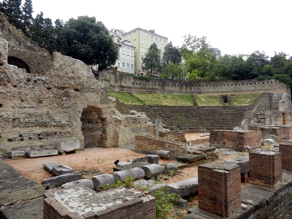 Column drums lying in a row on red gravel among square brick piers, beside a large mass of rubble masonry pierced by an arched opening, with the seating and its upper wall in the background.