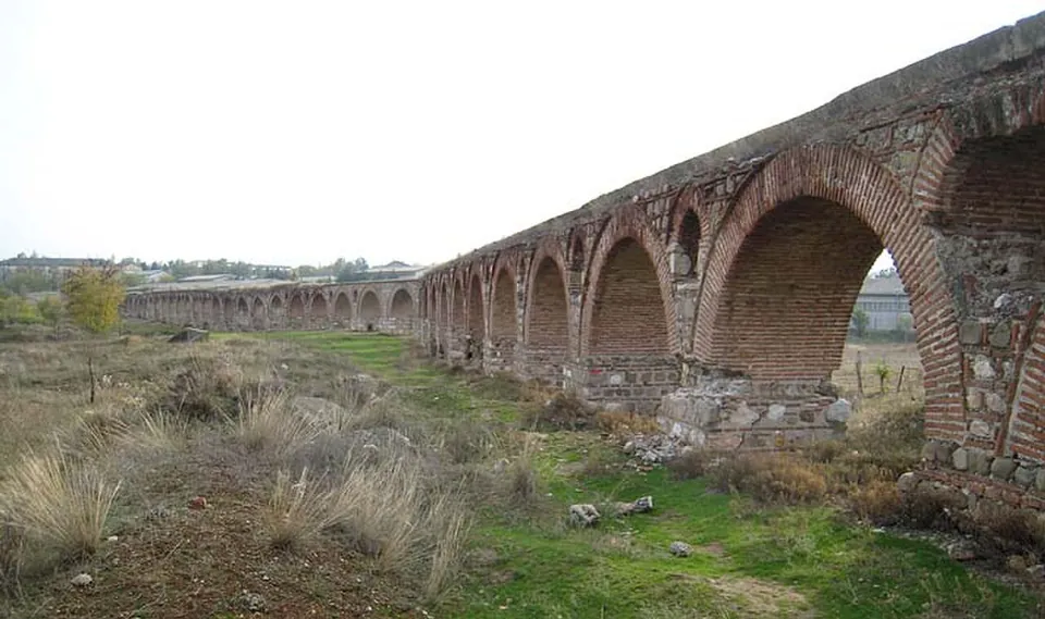 The arcade of brick arches receding into the distance across rough grassland under an overcast sky, with low buildings on the horizon.