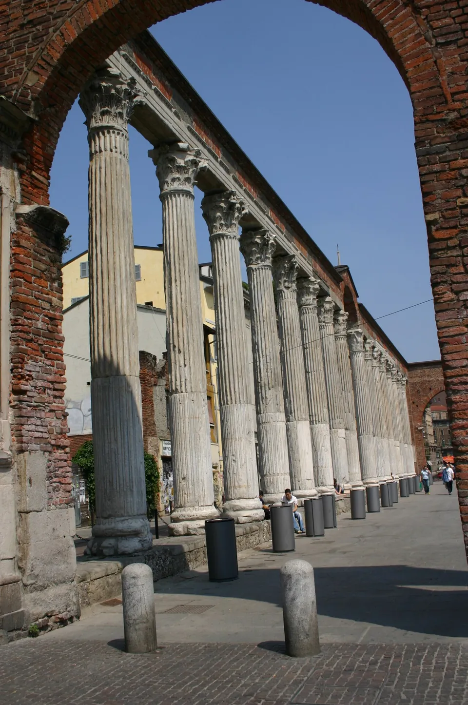 A long row of fluted columns with Corinthian capitals carrying a continuous entablature, seen along its length through a brick arch, with a paved street beside it.