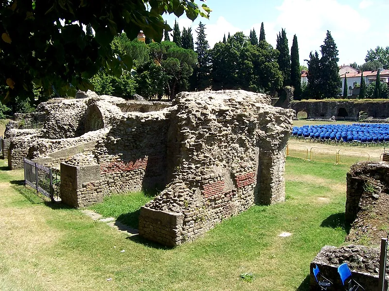 A broken block of rubble masonry laced with courses of red brick, standing on the grass, with lower walls of the same build behind it and rows of blue chairs in the distance.