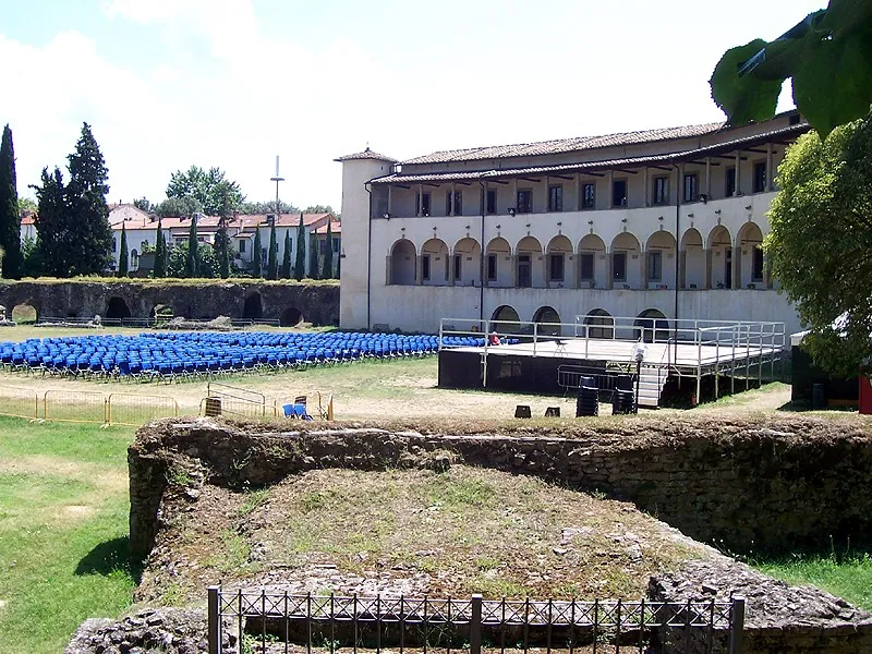 Rows of blue chairs and a raised stage set out on the arena floor in front of the arcaded museum building, with a low ruined wall in the foreground.