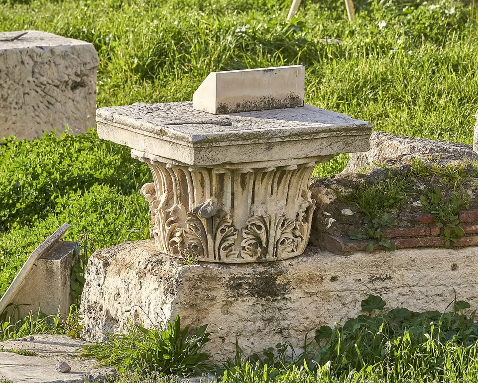 A marble column capital carved with curling acanthus leaves under a square slab, set on a rough stone block in the grass among other fallen blocks.