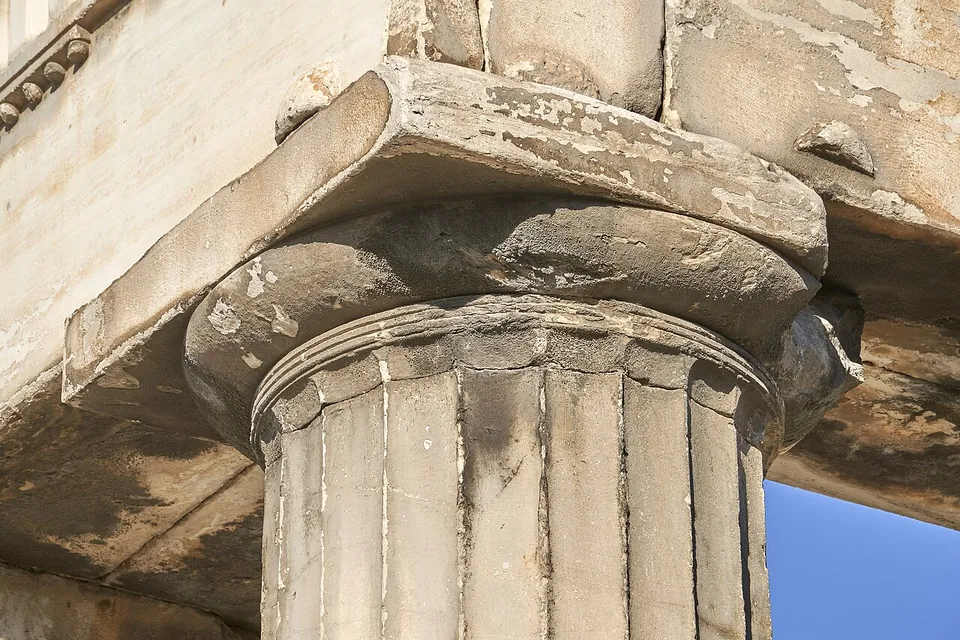 Close-up of the top of a fluted marble column, its plain cushion-shaped capital and square slab carrying a weathered beam against a blue sky.
