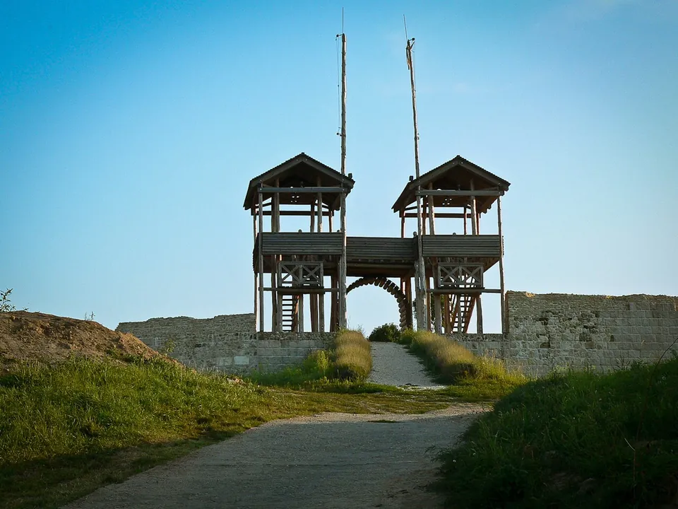 Two square timber towers joined by a wooden walkway standing over an arched gateway in a stone wall, with a path leading up to it between grassy banks under a blue sky.