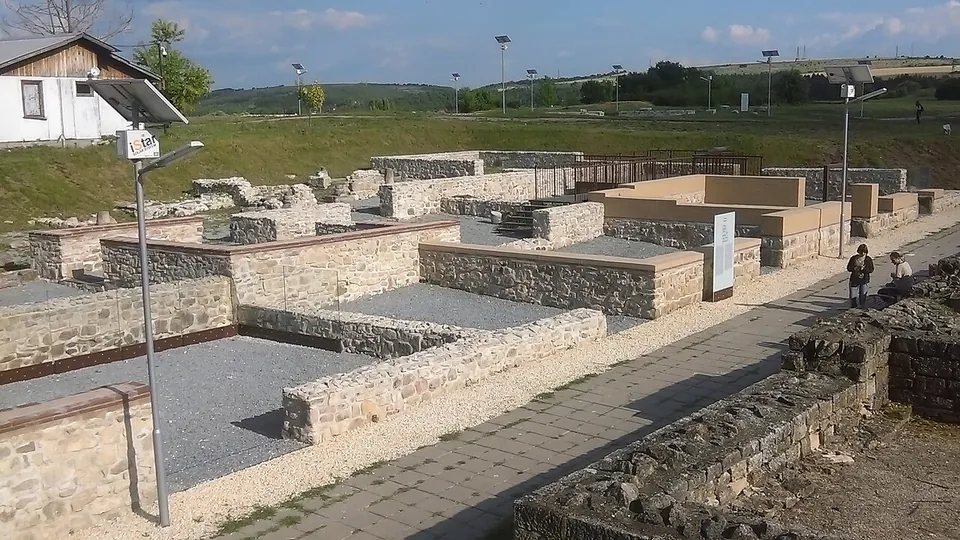 Restored low walls of pale stone outline rooms filled with grey gravel, crossed by a paved path with lamp posts and information panels, with grassy slopes and buildings behind.