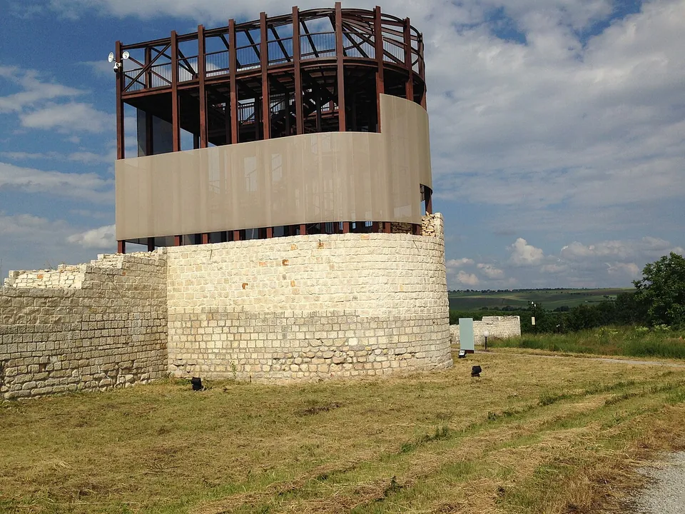 A round tower of pale squared stone rising from a stretch of wall on a mown slope, topped by a modern viewing platform of rust-red steel with a screen around its lower part.