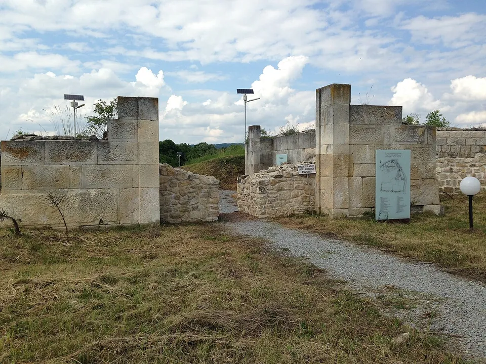 A gravel path runs between two restored gateway piers of large squared stones, with lower rubble walls inside and an information panel with a plan of the town beside the gate.