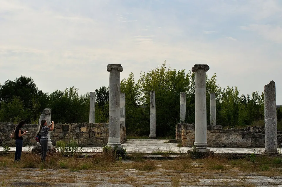Marble columns with Ionic capitals and broken column shafts stand around a paved open space, enclosed by low restored stone walls, with two visitors taking photographs and trees behind.