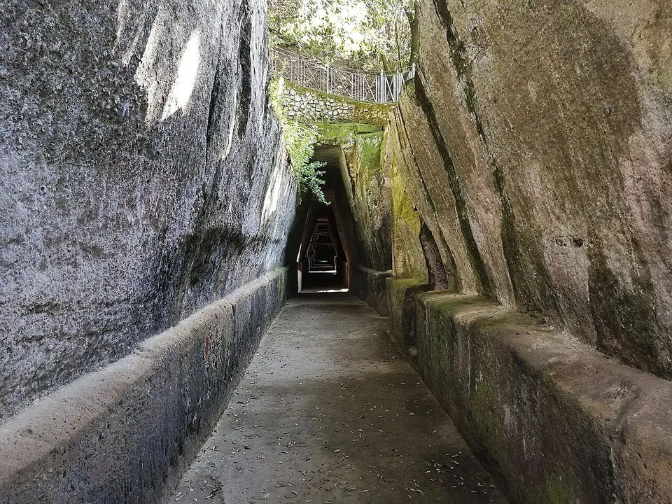 A long, narrow passage cut into pale tufa rock, its walls leaning inwards towards the top, running straight into darkness with a paved floor and a low stone kerb on each side.