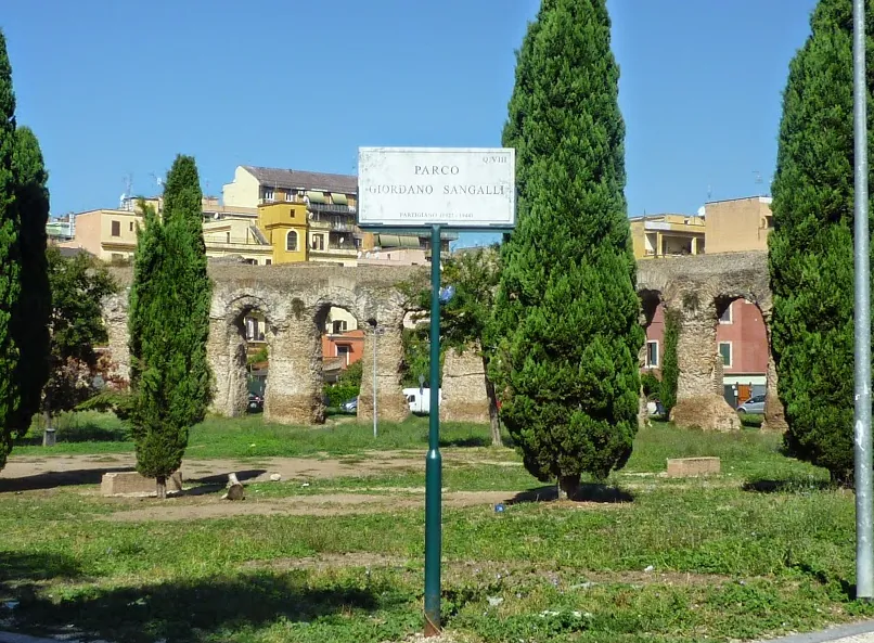 A street sign reading Parco Giordano Sangalli stands on a lawn between cypress trees, with a row of the aqueduct's arches and houses behind.