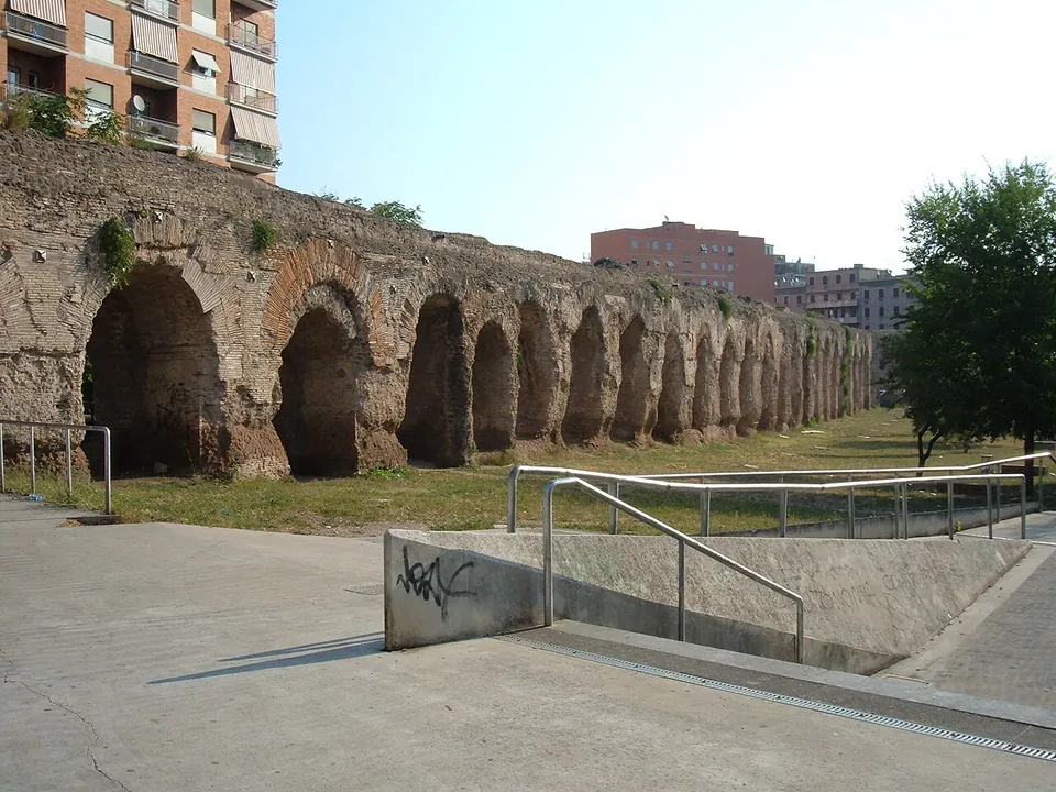 A long arcade of eroded stone and brick piers runs across a lawn beside a concrete ramp with metal railings, with apartment blocks rising behind it.
