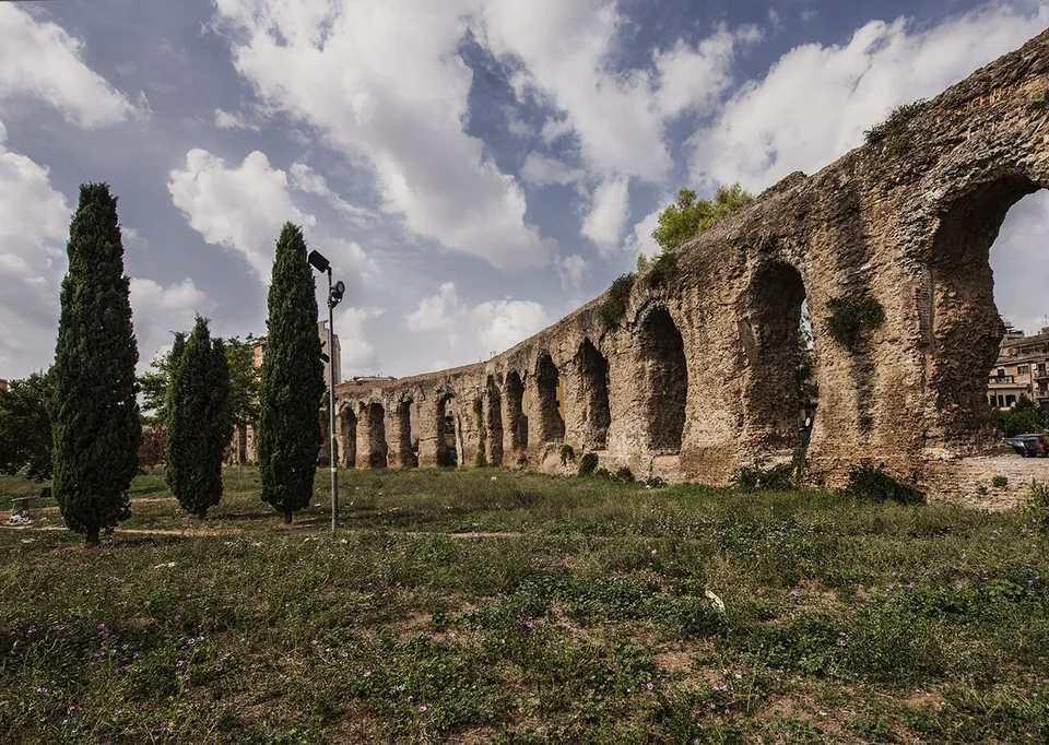 A long row of tall arches of weathered brick and stone curves away across a lawn, with cypress trees and a lamp post in front and apartment blocks just visible behind.