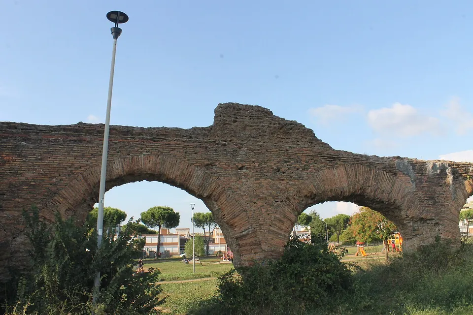 Two round arches built of red brick, the brick facing worn away in patches to the rubble core above them, with a park seen through the openings.