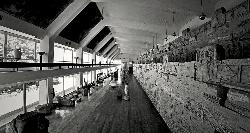 A black-and-white view down a long gallery with a timber floor and glazed wall, where carved stone panels with figures are set in rows along the opposite wall.