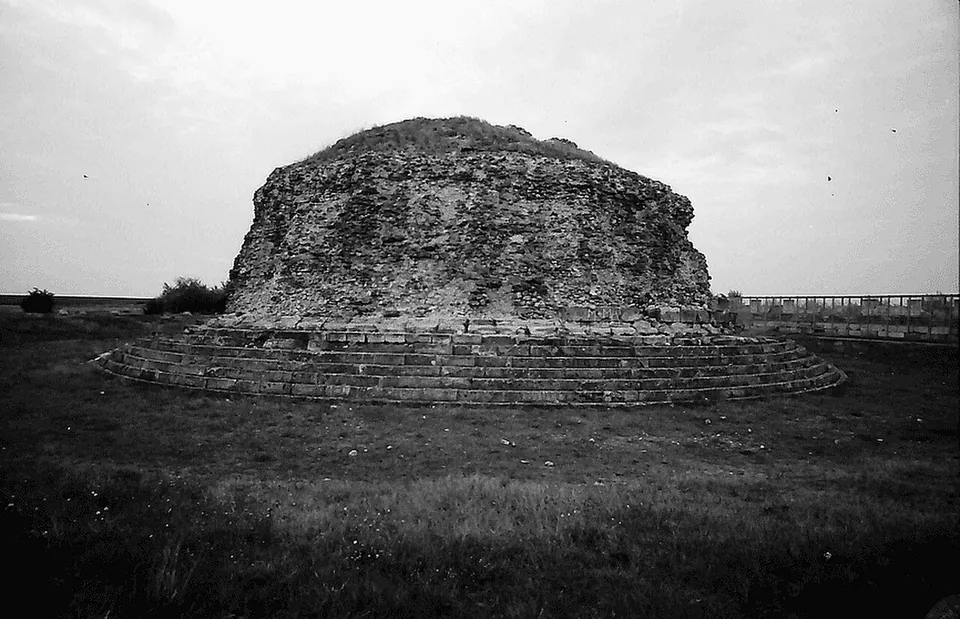 A black-and-white photograph of a low rounded mound of rough masonry rising from a ring of stone steps on open ground.