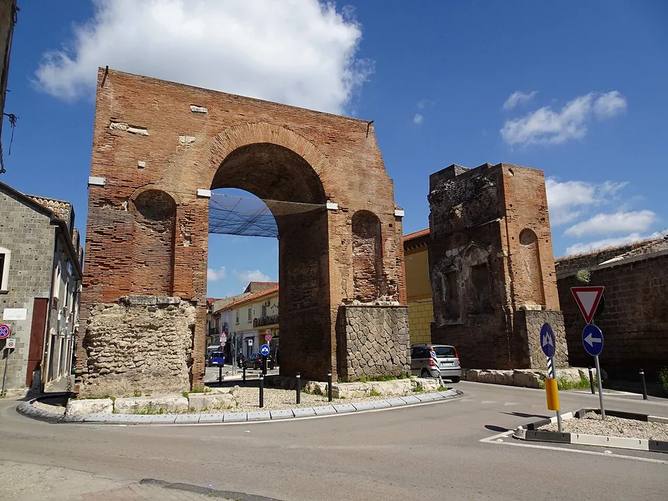 A tall brick arch on a base of stone blocks standing on a small traffic island at a road junction, with a separate brick pier with niches to its right and parked cars and houses around it.