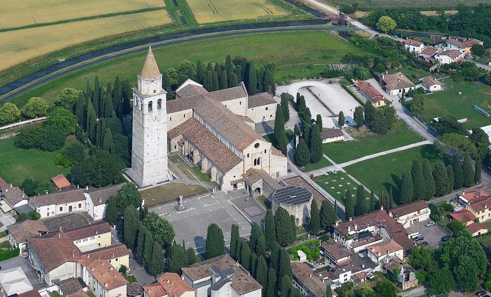 An aerial view of a large church with a tall square bell tower, surrounded by red-roofed houses, lawns and lines of cypresses, with fields and a canal beyond.