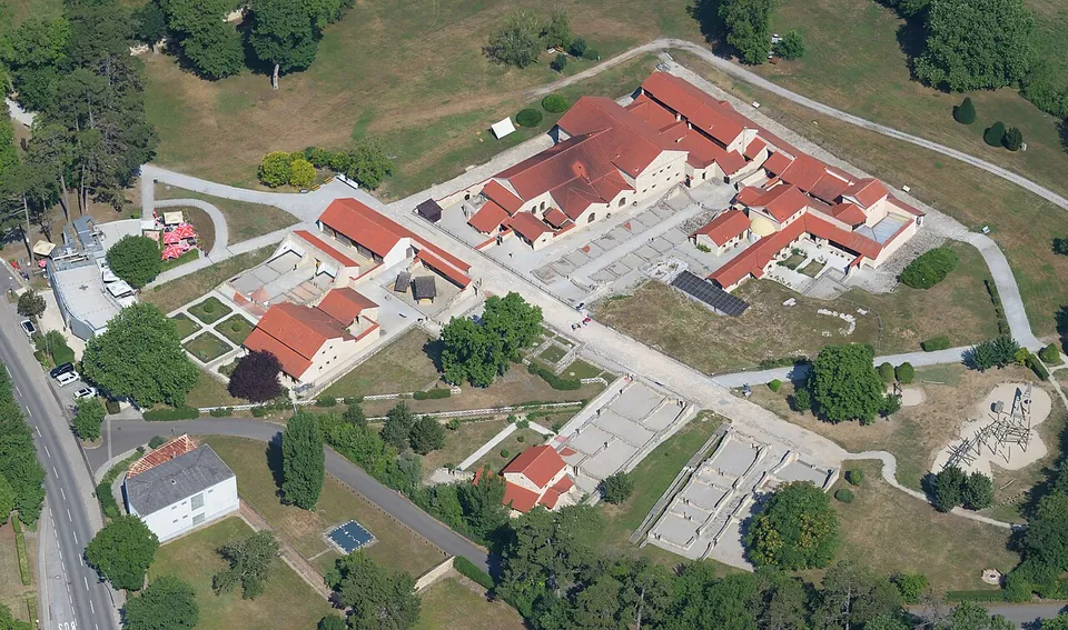 An aerial view of a cluster of rebuilt Roman houses with red tiled roofs around courtyards, next to excavated wall foundations in grass, with paths and trees around them.