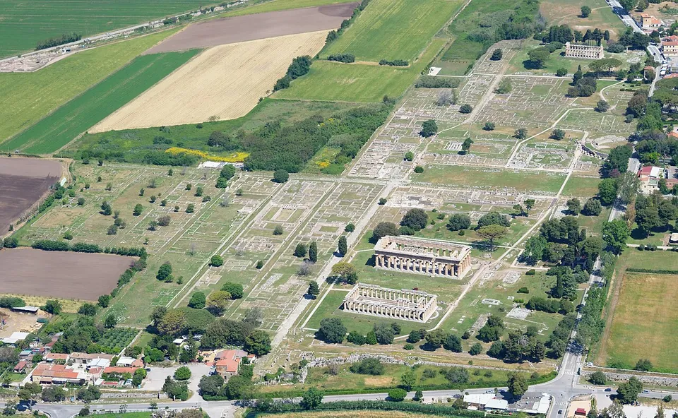 An aerial view over farmland of a long grid of low excavated walls and streets in mown grass, with two large columned temples standing side by side in the foreground and the modern village along the bottom edge.