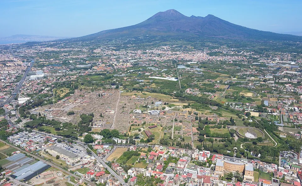 An aerial view over the modern town to the excavated city, a wide grid of roofless grey-brown blocks set among green fields and trees, with the cone of Vesuvius rising on the horizon.