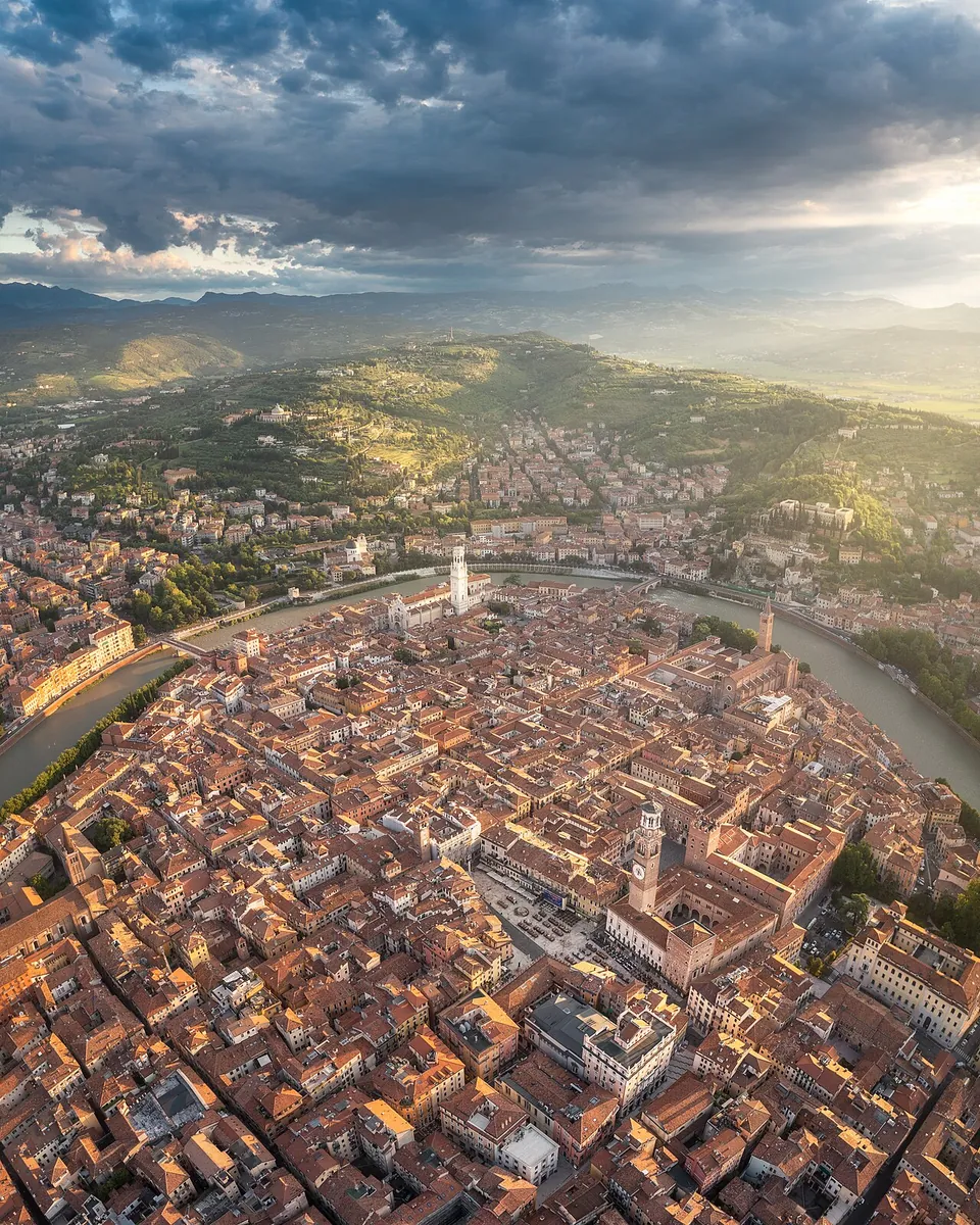 An aerial view at dawn over a dense city of red-tiled roofs inside a tight loop of a river, with bell towers and a large square, wooded hills rising behind and mountains on the horizon.
