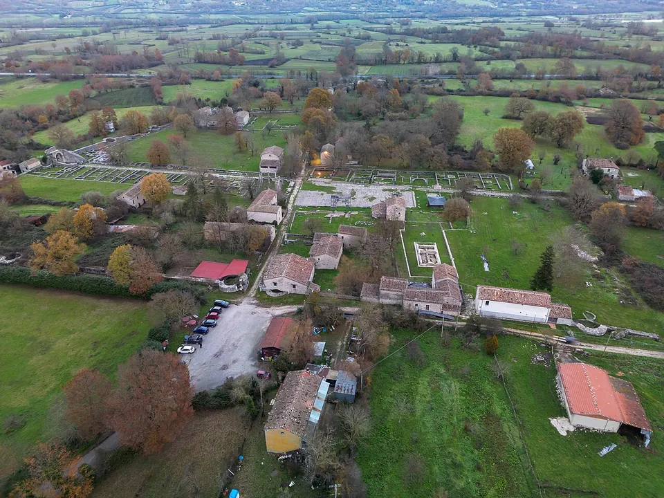 An aerial view over green fields and scattered trees, with a cluster of stone farmhouses with red-tiled roofs, a gravel car park and the low outlines of excavated buildings in the grass between them, and rolling farmland running to the hills behind.