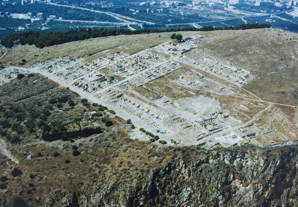 An aerial view of a hillside plateau above a rocky cliff, covered with the low walls of a town laid out in regular rectangular blocks along straight streets, with trees and modern buildings in the plain beyond.