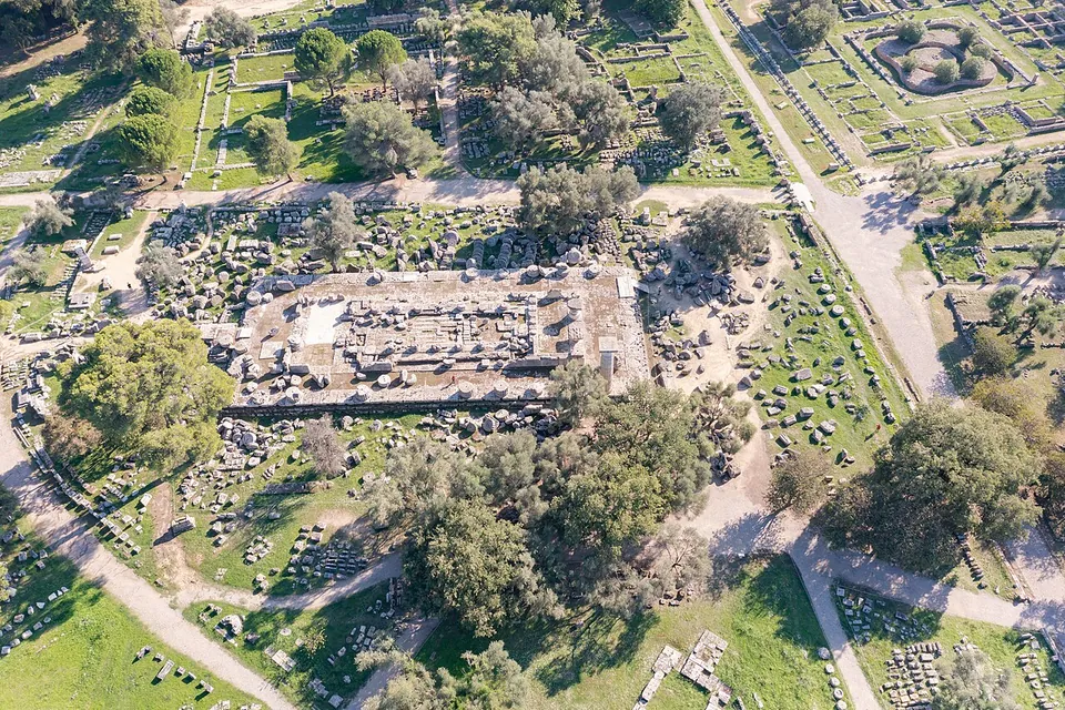 The sanctuary seen from the air, the long rectangular platform of a great temple strewn with fallen column drums at the centre, surrounded by trees, foundations and gravel paths through green grass.