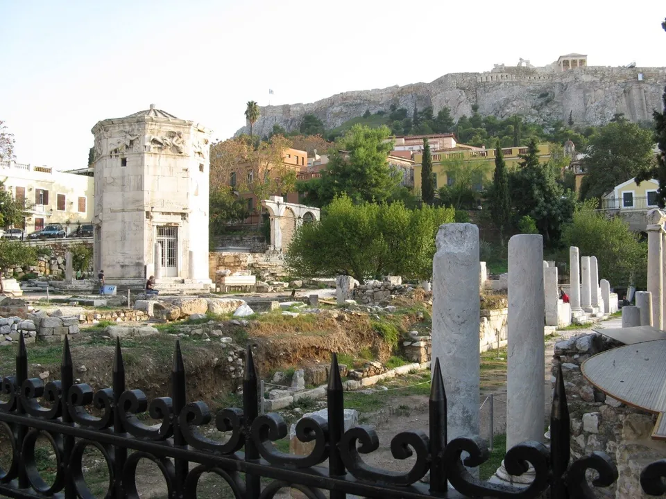 Looking over an iron railing across excavated ground to an octagonal marble tower, a ruined arched gateway and standing column shafts, with the Acropolis rock and its temples high behind.
