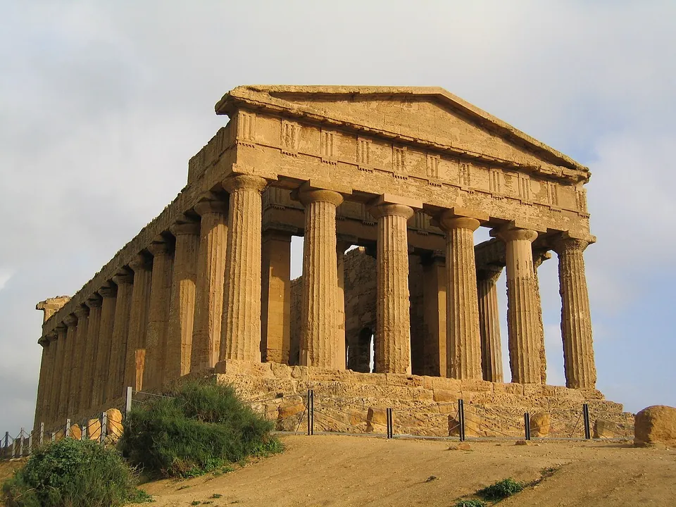 A Doric temple seen from one corner, its columns, entablature and front pediment all standing on a stepped base, built of warm golden stone, with dry ground and shrubs in front.