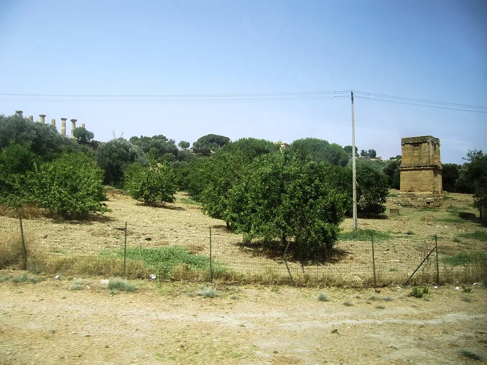 A tall square stone monument standing alone among fruit trees and dry grass behind a wire fence, with a row of columns on the ridge in the distance at the left.