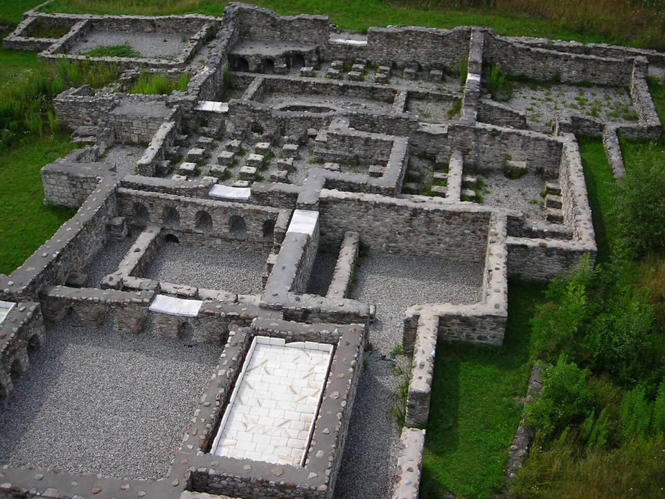 The excavated walls of a bath building seen from above, rooms of grey stone with rows of small square pillars that held up heated floors, and a pale stone slab in one room.
