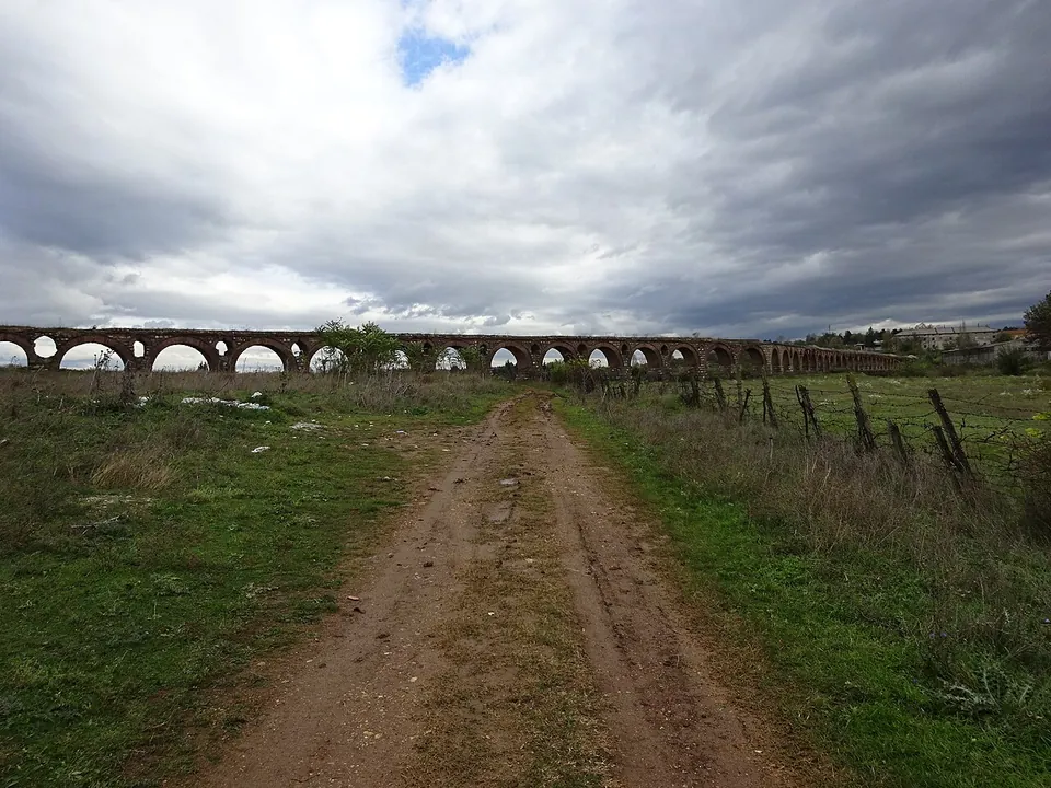 A muddy dirt track between fields leading towards the long line of the aqueduct on the horizon, under a heavy cloudy sky, with a wire fence on the right.