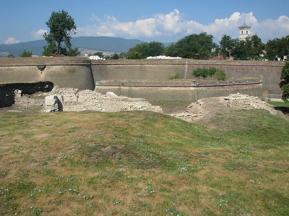 Grassy mounds and low ruined walls in the foreground, with the long sloping brick-and-stone rampart of a star-shaped fortress behind and a church tower beyond the trees.